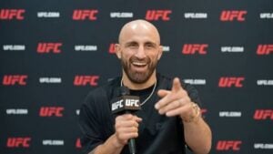 UFC featherweight champion Alexander Volkanovski smiles and points while holding a UFC microphone in front of a UFC backdrop