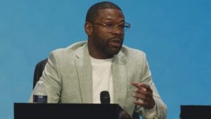 Floyd Mayweather in glasses and a light green blazer speaks at a table with a microphone in front of a blue backdrop