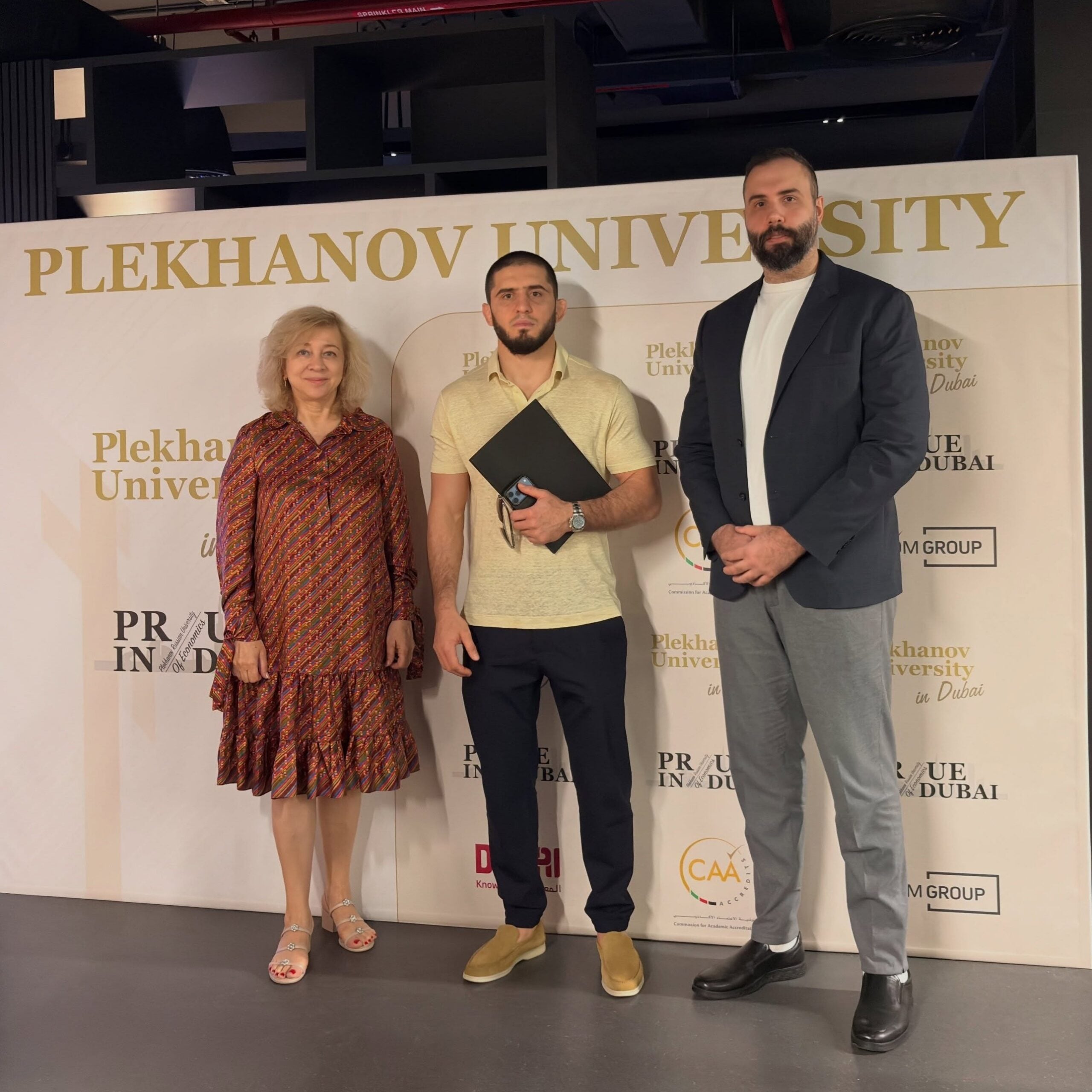 Islam Makhachev poses with two Plekhanov University representatives in front of a Plekhanov University in Dubai backdrop